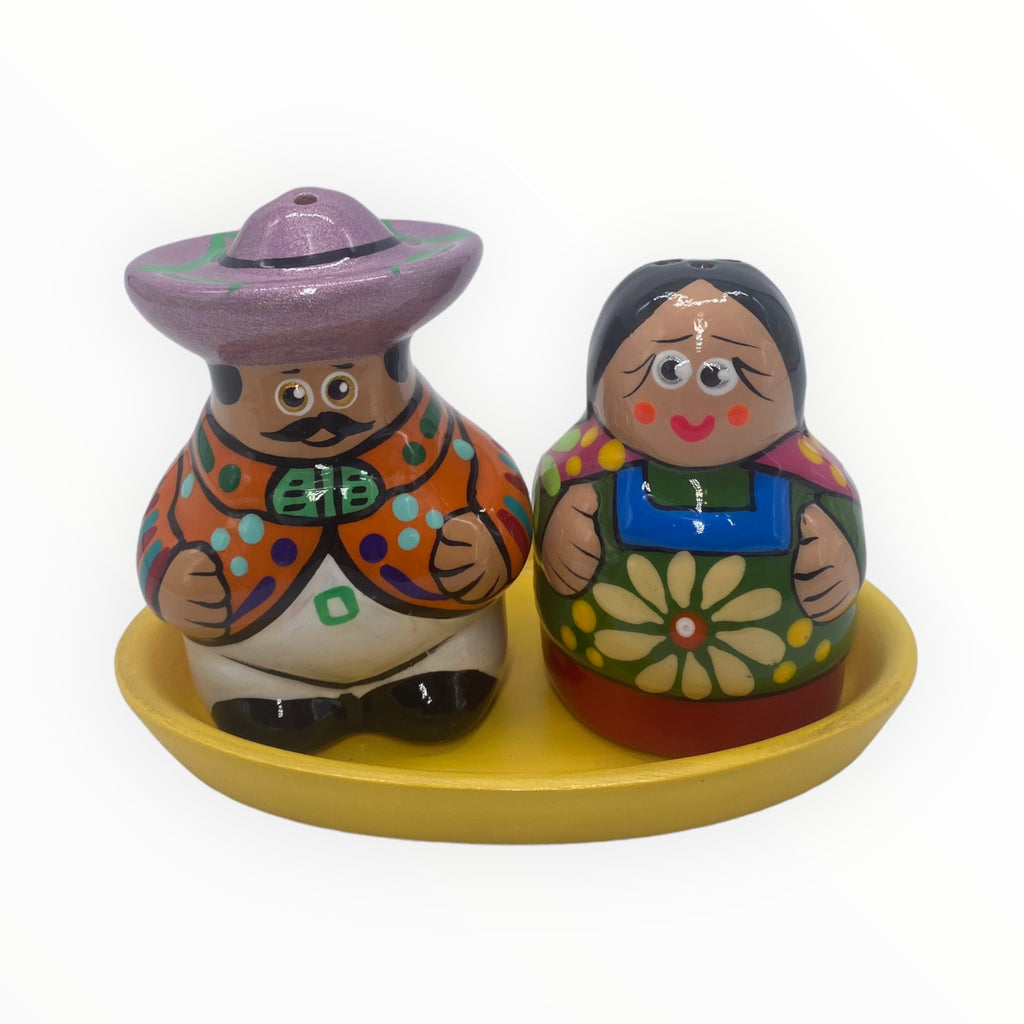 "Experience the beauty of Mexican craftsmanship with this one-of-a-kind salt and pepper shaker pair. The gentleman dons a stylish mustache, an eye-catching lilac hat, and a charming orange jacket, while the lady captivates with her sweet expression and a green dress featuring a delicate flowers accent. 